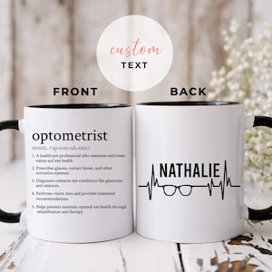 Op de afbeelding: Witte keramische koffiemok met een zwarte rand en handvat. De voorkant van de mok heeft de definitie van het woord "optometrist" in zwart gedrukt. De achterkant van de mok heeft een zwarte hartslaggrafiek met een bril en de naam "Nathalie" in zwart gedruckt.