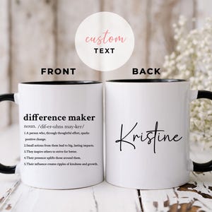 May include: White ceramic coffee mug with a black rim and handle. The front of the mug has the text "difference maker" with a definition. The back of the mug has the name "Kristine" in black cursive.