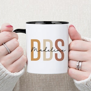 May include: White ceramic mug with a black rim and handle. The mug features the letters "DDS" in shades of brown, with the name "Madeline" in a script font. The mug is held by a person wearing a cream-coloured sweater.