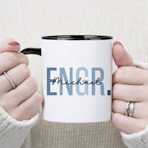 May include: White ceramic mug with a black handle and rim. The mug features the word "ENGR" in blue block letters, with the name "Michael" in a script font underneath. A period follows the name.