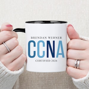 May include: White ceramic mug with a black rim and handle, featuring the name "Brendan Werner" and the letters "CCNA" in blue, with "CERTIFIED 2026" below. The mug is held by hands wearing rings.