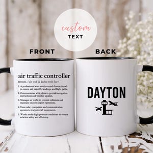 May include: White ceramic mug with a black rim and handle. The front of the mug has the definition of an air traffic controller. The back of the mug has the word "DAYTON" and a graphic of an airport control tower with airplanes.