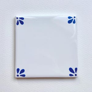 May include: Square white ceramic tile with glossy finish. Each corner features a blue floral design. The tile is a decorative element, suitable for various home decor projects. The tile's clean design and color scheme make it versatile.