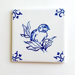 May include: A square white ceramic tile featuring a blue ink-style illustration of a small bird perched on a leafy branch. The corners of the tile are decorated with blue floral scrollwork motifs, creating a symmetrical and clean visual design.