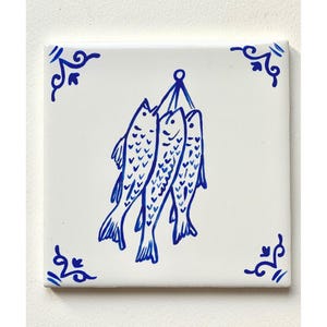 May include: A white ceramic tile with a blue and white design of three fish hanging from a string. The tile has a decorative border with blue swirls.