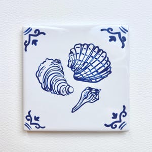 May include: Square white ceramic tile featuring blue seashell illustrations. The tile has three different seashell designs and decorative blue corner accents. A coastal or nautical theme.