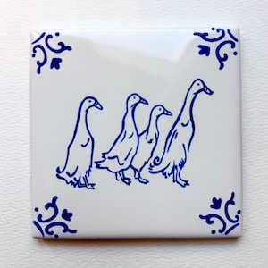 May include: White square ceramic tile featuring a blue illustration of four geese walking in a row. The tile has decorative blue corner accents. The design is simple and elegant, suitable for home decor.