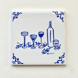 May include: Blue and white ceramic tile with a hand-drawn illustration of a bottle of wine, four glasses, and figs. The tile has a decorative border.