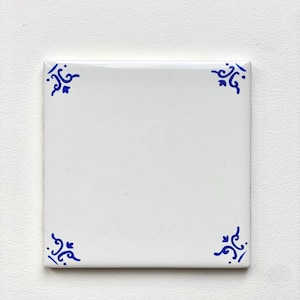 May include: Square white ceramic tile with blue decorative corner designs. The tile has a glossy finish and a simple, clean aesthetic. The blue designs are symmetrical and add a touch of elegance to the tile's design.