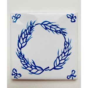 May include: A white ceramic tile with a blue wheat design. The wheat is arranged in a circle around the center of the tile. There are four small blue floral designs in the corners of the tile.