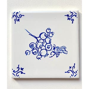 May include: A white ceramic tile with a blue and white design of a bee and a cluster of berries. The tile has a decorative border.
