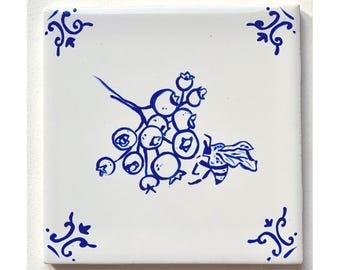 May include: A white ceramic tile with a blue and white design of a bee and a cluster of berries. The tile has a decorative border.