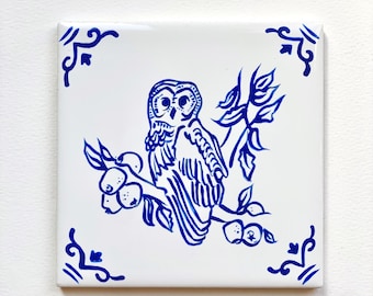 May include: A blue and white ceramic tile with a decorative design of an owl perched on a branch with apples. The owl has a detailed design with feathers and a round eye. The tile has a blue and white border with a decorative design.