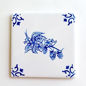 May include: A square ceramic tile with a white base and a hand-painted blue design. The center features a detailed illustration of a blackberry branch with leaves and fruit. Each of the four corners is decorated with a small blue scroll motif.