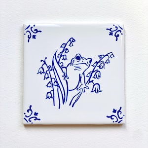 May include: A blue and white ceramic tile with a hand-painted design of a frog sitting on a lily of the valley plant.