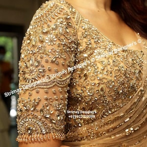 May include: A gold-toned blouse featuring intricate embroidery with clear crystals, sequins, and metallic thread work on sheer fabric. The garment includes a scalloped sleeve edge with dangling bead accents and the text Strimay Designer's.