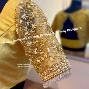 Puede incluir: Primer plano de una manga de blusa dorada con intrincados bordados. La manga presenta una superposición transparente adornada con hilo dorado, perlas y motivos florales. El texto "Strimay Designer's" es visible. La manga tiene un fleco de cuentas.