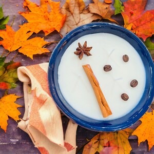 Könnte beinhalten: Eine weiße Kerze in einer blauen Keramikschale mit Zimtstange, Sternanis und Kaffeebohnen darauf. Die Kerze ist von Herbstblättern umgeben.