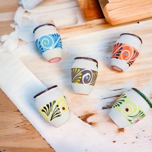 May include: Five small, white ceramic pots with colorful painted designs. The pots have a rounded shape and a small opening at the top. The designs include swirls, dots, and lines in shades of blue, yellow, orange, and brown.