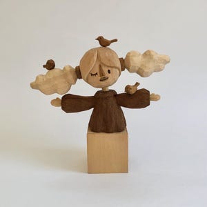 May include: A wooden figurine of a person with brown hair and a brown dress. The person has a winking eye and is holding out their arms. There are three small birds perched on the person's hair and arms.