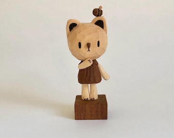 Puede incluir: Figura de oso de madera con una pequeña abeja en la cabeza. El oso es de color marrón claro con detalles en marrón oscuro y lleva un vestido marrón. Se encuentra sobre una base cuadrada de madera.