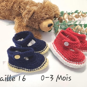 Peut inclure: Deux paires de chaussons pour bébé tricotés. Une paire est bleu marine avec une semelle jaune et un bouton en bois. L'autre paire est rouge avec une semelle blanche et un bouton blanc. Un ours en peluche est en arrière-plan.