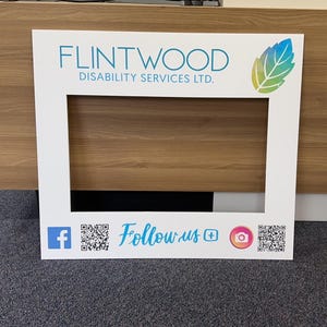 May include: White photo booth frame with the text "FLINTWOOD DISABILITY SERVICES LTD." and a green leaf design. The frame has a cutout for a person's head and includes social media icons and QR codes for Facebook and Instagram.