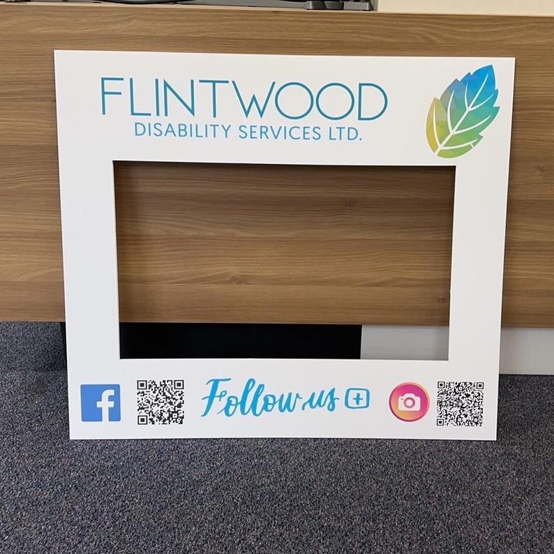 May include: White photo booth frame with the text "FLINTWOOD DISABILITY SERVICES LTD." and a green leaf design. The frame has a cutout for a person's head and includes social media icons and QR codes for Facebook and Instagram.