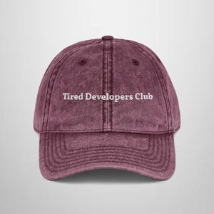 Puede incluir: Gorra de béisbol granate con el texto "Tired Developers Club" en blanco. La gorra tiene una visera curva y un diseño clásico de seis paneles. La tela tiene un aspecto vintage ligeramente descolorido.