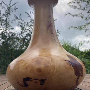 Könnte beinhalten: Eine gedrechselte Holzvase mit glatter, polierter Oberfläche, die einen breiten, abgerundeten Boden besitzt, der sich zu einem schlanken Hals und einem ausgestellten Rand verjüngt. Das helle Holz zeigt natürliche Maserungen und dunkle Knoten.