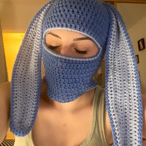 May include: A blue crochet balaclava with long bunny ears. The face opening is outlined in a lighter blue. The handmade head covering is a unique accessory.