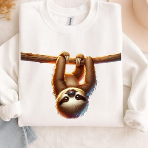 Puede incluir: Sudadera blanca con un perezoso marrón colgando de una rama. El perezoso tiene pelaje marrón y una cara sonriente. La rama es marrón, y la sudadera es lisa.