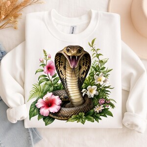 May include: A white crewneck sweatshirt featuring a central graphic of a brown cobra with its hood flared and mouth open, surrounded by vibrant pink hibiscus flowers, white blossoms, and lush green tropical foliage and fern leaves.
