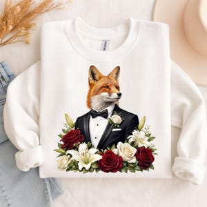 Puede incluir: Una sudadera blanca de cuello redondo con un estampado gráfico de un zorro vestido con esmoquin negro, camisa blanca, pajarita negra y un boutonniere de rosa blanca, rodeado por un arreglo floral de rosas rojas y lirios blancos.