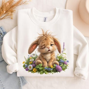 May include: A white long-sleeve sweatshirt featuring a graphic of a fluffy brown bunny with messy fur winking while sitting in a patch of colorful wildflowers including daisies, purple clover, and yellow blossoms on a green grassy base.
