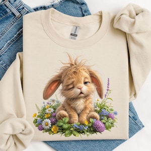 May include: A beige crewneck sweatshirt featuring a graphic of a fluffy light brown bunny with messy fur winking while sitting in a patch of colorful wildflowers including daisies, purple clover, and green leaves. The garment has a Gildan brand label.