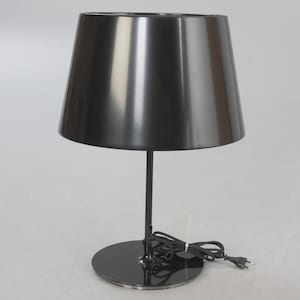 May include: A black table lamp with a conical shade and a circular base. The lamp has a slender stem and a power cord with a plug. The shade is angled and reflects light. The lamp is a modern design.