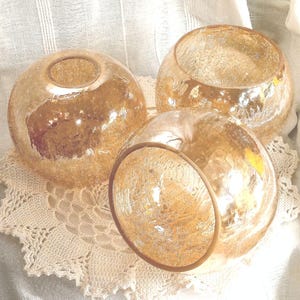 Könnte beinhalten: Drei kugelförmige, goldfarbene Kerzenhalter aus Glas mit strukturierter Oberfläche. Die Kerzenhalter sind auf einer cremefarbenen Spitzendecke angeordnet. Die offenen Oberseiten der Kerzenhalter sind sichtbar.