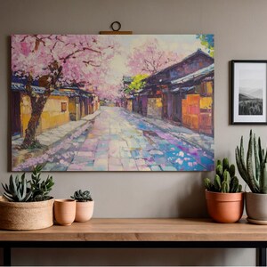 以下が含まれることがあります： 満開の桜が咲く日本の街並みの油絵。伝統的な建物が並ぶ石畳の通りを描いています。絵画は、植木鉢に入った植物が置かれた木製のテーブルの上にある壁に掛けられています。