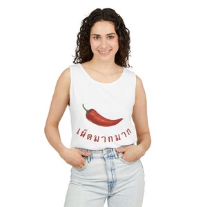 May include: A white sleeveless tank top featuring a graphic of a single red chilli pepper centred on the chest. Below the chilli pepper, the Thai text "เผ็ดมากมาก" is printed in a dark red font. The garment has a round neckline and wide armholes.