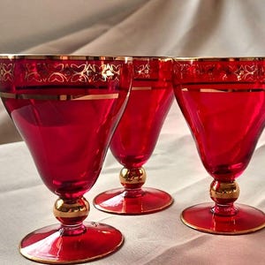 Puede incluir: Tres copas de vino rojo rubí con bordes dorados y detalles dorados decorativos. Las copas tienen una base redonda y una bola dorada en el tallo. Las copas están dispuestas sobre una superficie blanca.