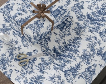Puede incluir: Un regalo envuelto y papel de regalo con un diseño de toile de Jouy azul y blanco con figuras de Papá Noel y árboles de hoja perenne. El regalo está atado con una cinta marrón. Tijeras doradas están sobre el papel.