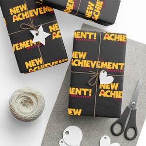 May include: Black wrapping paper featuring the repeating yellow text NEW ACHIEVEMENT! with small red pixelated heart icons. The paper is tied with brown twine and includes white star and heart-shaped gift tags, a ball of yarn, and scissors.