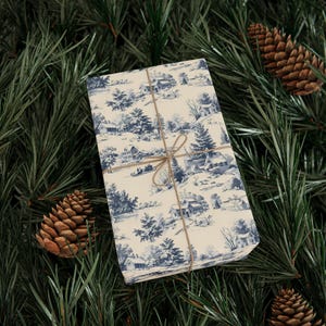 A rectangular box wrapped in blue and white winter village paper and tied with twine rests amidst green pine branches and three pinecones.