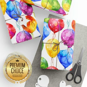 Two packages wrapped in colorful watercolor balloon paper with twine, sitting on a gray surface next to black scissors, tags, and a gold premium choice seal.