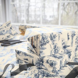 Two gift boxes wrapped in blue and white winter village paper sit with black ribbon and scissors against a window with a blurred view of trees.