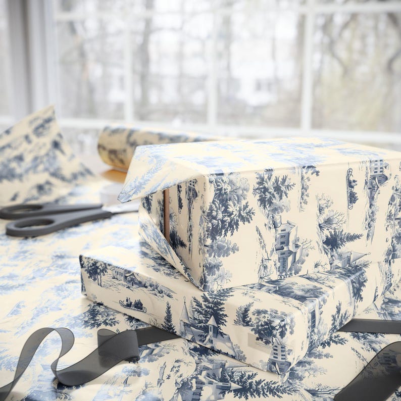 Two gift boxes wrapped in blue and white winter village paper sit with black ribbon and scissors against a window with a blurred view of trees.