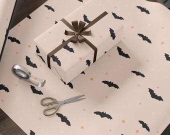 Op de afbeelding: Een ingepakte geschenkdoos met een bruin lint en strik ligt op een vel cadeaupapier. Het papier en de doos zijn lichtroze met een patroon van zwarte vleermuizen en kleine oranje en witte sterren. Schaar en plakband.