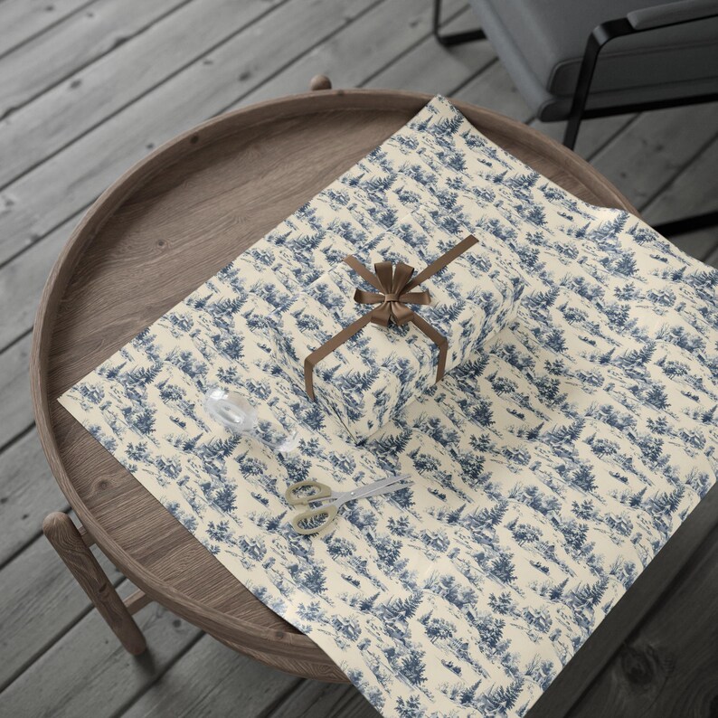 A small wrapped gift tied with a brown ribbon sits on a sheet of blue and white winter village wrapping paper atop a round wooden tray on a slatted wood surface.