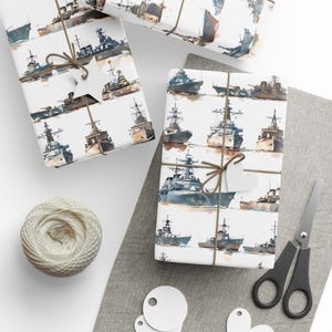 A wrapped gift box featuring watercolor warships patterned on white paper, placed on a gray mat next to a ball of twine, scissors, and blank tags.